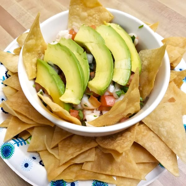 A white bowl of ceviche with sliced avocado, surrounded by tortilla chips on a patterned paper plate.