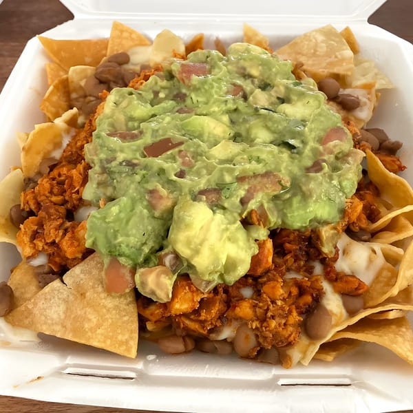 Mexican nachos with chicken, beans, cheese, and a large dollop of guacamole in a white takeout container.